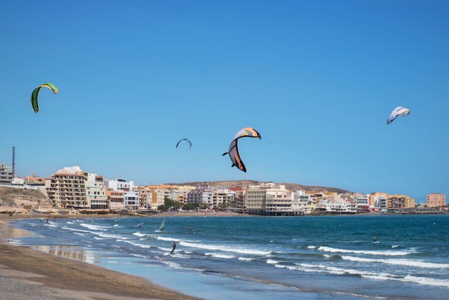 Kitesurfers maneuvering on the waves at El Médano Beach, Tenerife, with colorful kites against a clear blue sky.