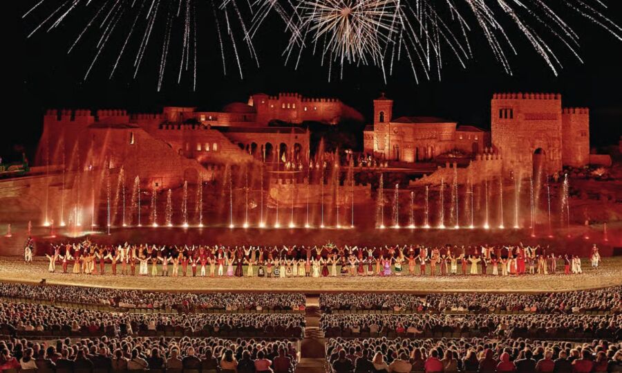 El Sueno de Toledo Show vibrant outdoor performance in front of a historic castle, with performers in colorful costumes and fountains lit up. Fireworks burst overhead, thrilling the large audience.