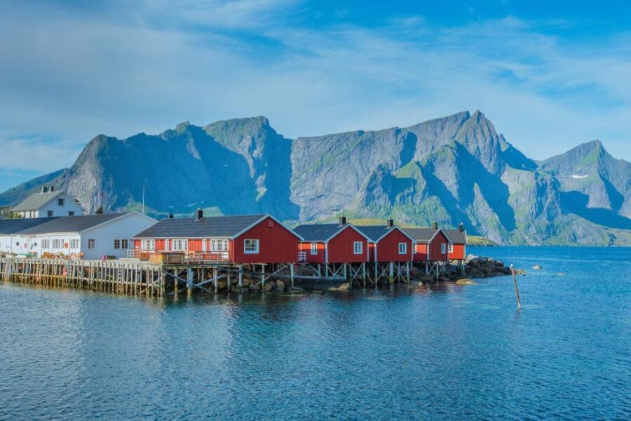 Eliassen Rorbuer and serene seaside village with red and white houses on stilts, set against majestic mountain peaks under a clear blue sky, evoking a peaceful, idyllic feel.