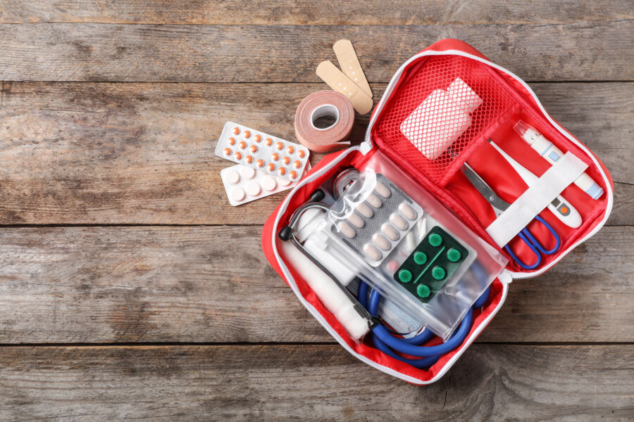 Open red first aid kit sits on a wooden surface, displaying bandages, scissors, a stethoscope, blister packs of pills, tape, and a white bottle.