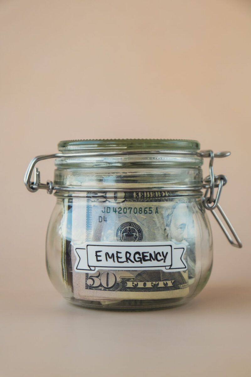Glass jar filled with US dollar bills labeled "Emergency" against a beige background, conveying preparedness and financial caution.