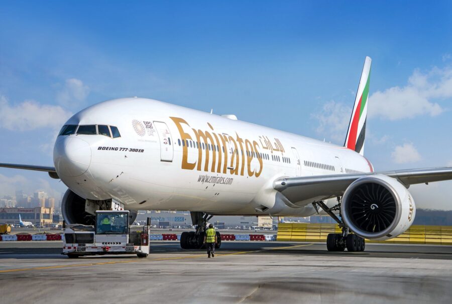 Emirates Boeing 777-300ER at airport with ground crew and vehicle, under clear skies.