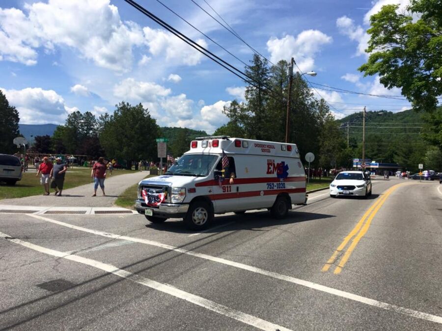 Ambulance at community event intersection with trees, police presence, and recreational activities.