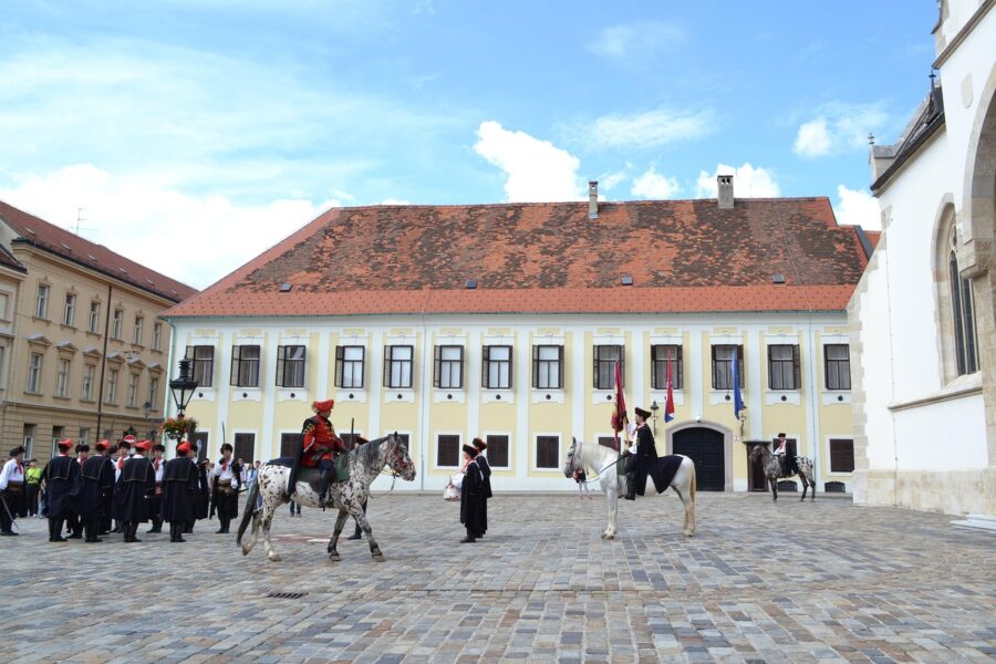 Ceremonial military parade in European courtyard with historic architecture and mounted figures.