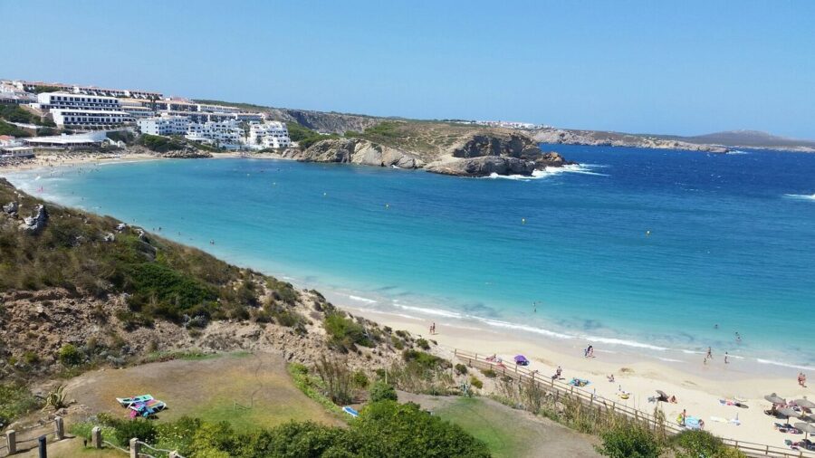 Es Mercadal, Menorca: a picturesque beach with a vibrant blue ocean and charming white buildings nearby.