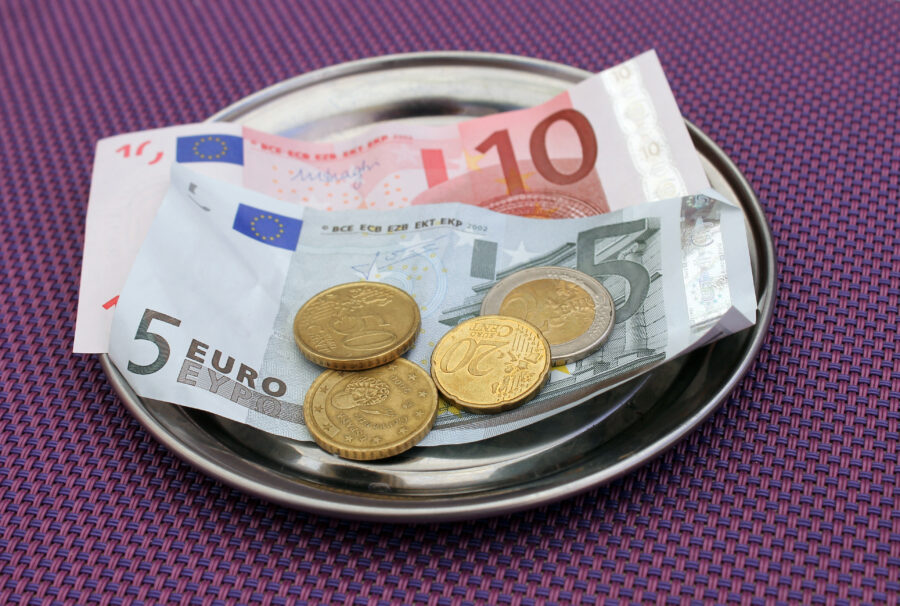 Small silver tray holds euro coins and notes, including 5 and 10 euro bills, on a purple woven placemat, conveying a restaurant tip.