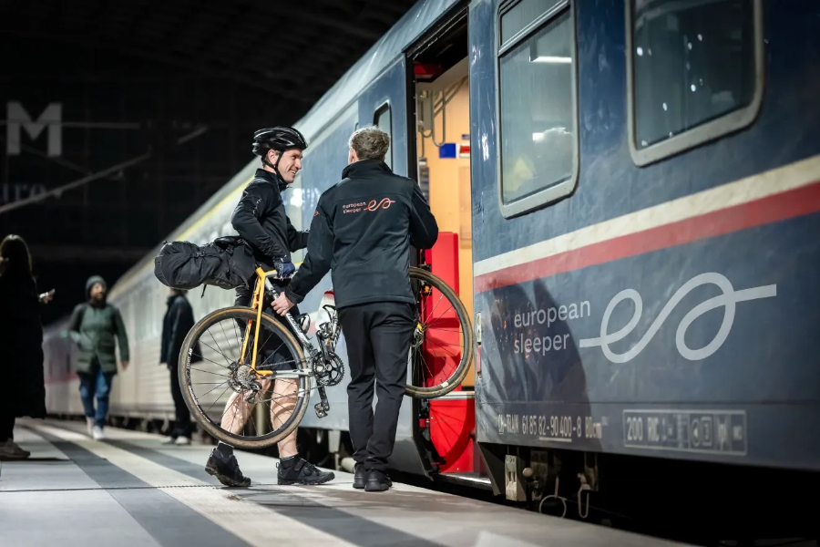 European sleeper train exterior with a woman entering while pushing her bike alongside the train.
