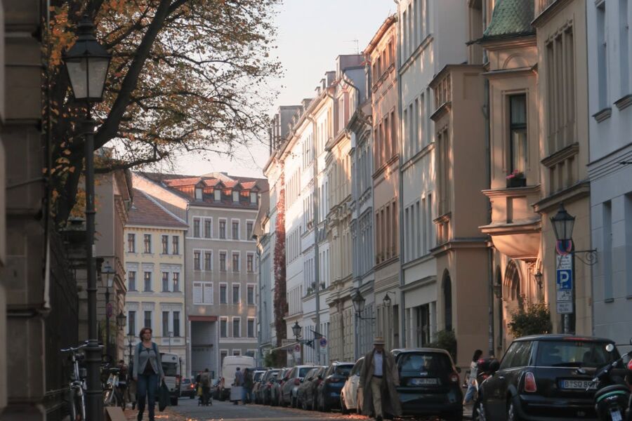 Serene European street with historical buildings, autumn trees, parked cars, and pedestrians in warm sunlight.