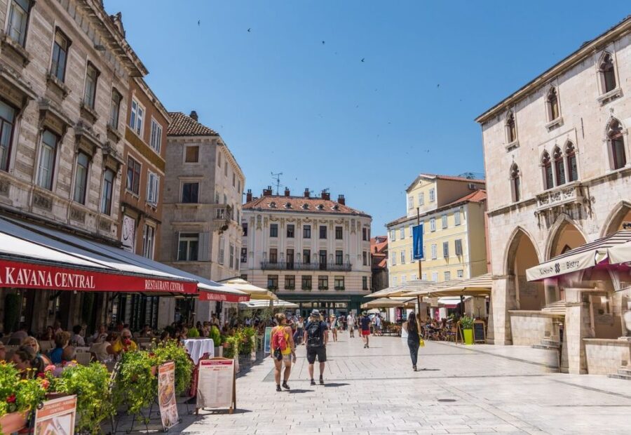 Vibrant Zagreb plaza, historical buildings, outdoor dining, diverse people, sunny atmosphere.