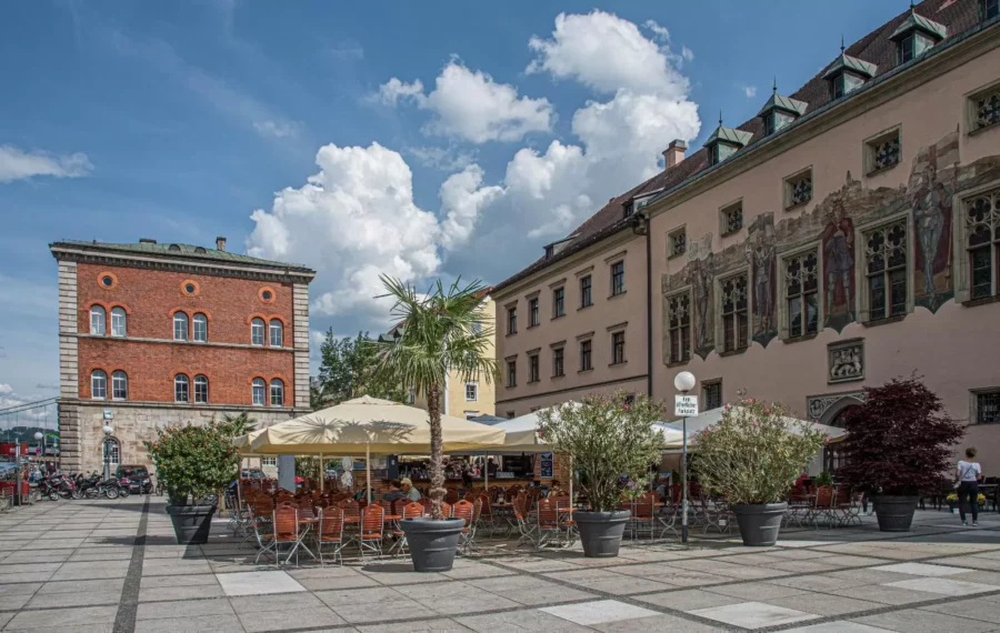 Sunny European old town square with outdoor café terrace, historic buildings, murals, and blue sky.
