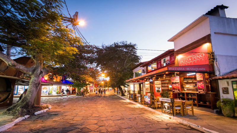 Dusk pedestrian street with pizza restaurant, shops, stone pavement, trees, warm lights and tourists.