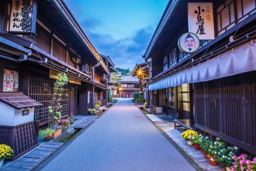 Quiet traditional Japanese street at twilight with wooden houses, lanterns, flowers, and mountain backdrop.