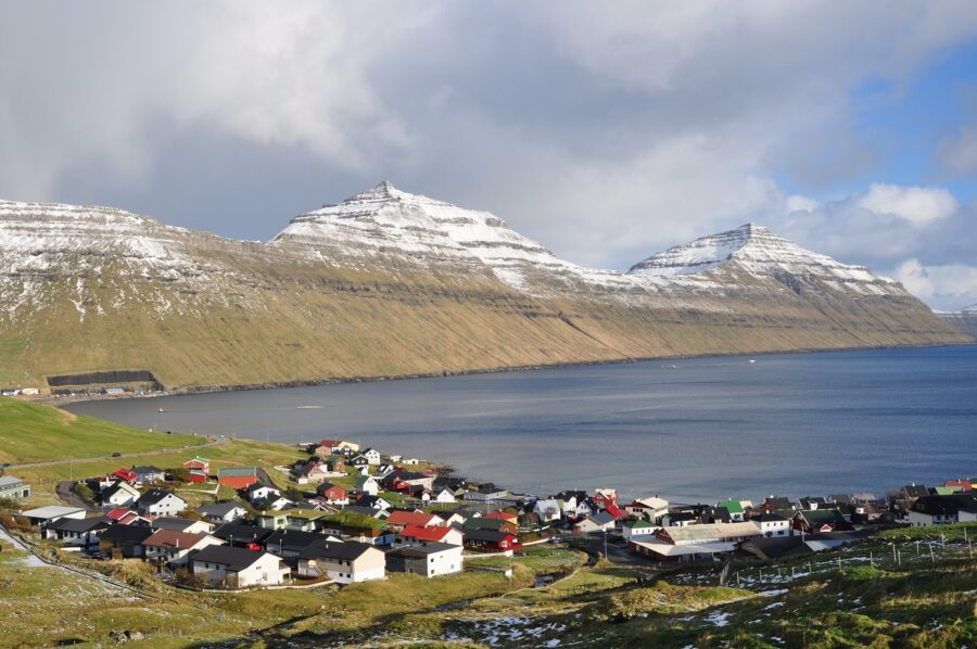 Scenic view of a small town in Eysturoy, Faroe Islands, with snow-capped mountains and a tranquil lake in the foreground.