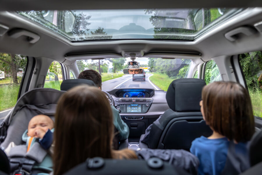Interior view of a car with three children in the back seat, one sleeping in a car seat. The driver is focused, driving on a scenic road.
