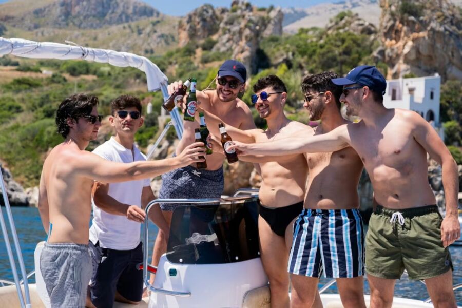 Group of six men on a boat joyfully toasting with beer bottles under a sunny sky. They're in swimwear, surrounded by mountains and blue waters.