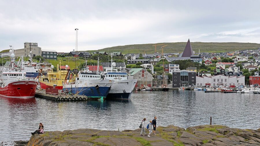 People gather on a rock near the harbor in Tórshavn, Faroe Islands, with scenic views of the water and boats.