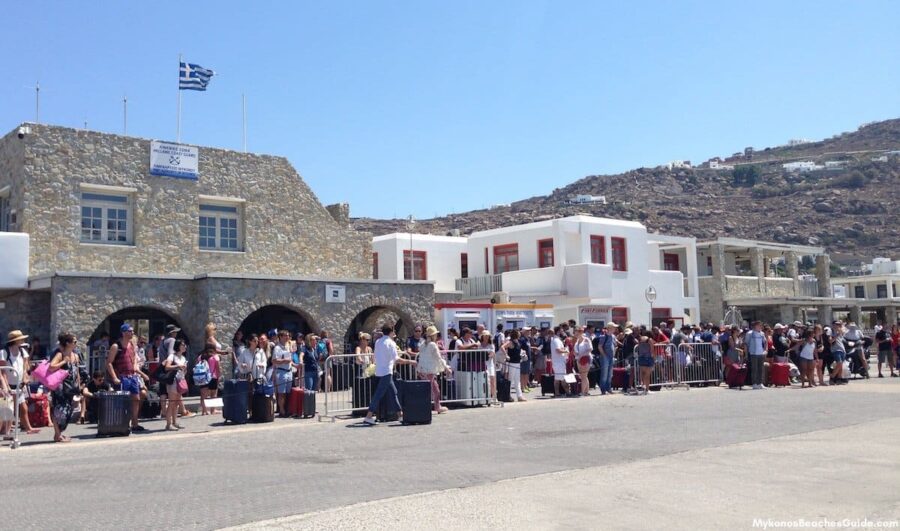 Busy Greek ferry terminal with travelers, luggage, vibrant architecture, and picturesque landscape.