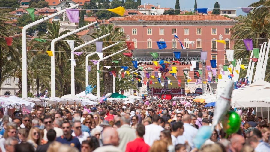 Lively festival promenade with palm trees, colorful decorations, diverse crowd, and vibrant coastal backdrop.