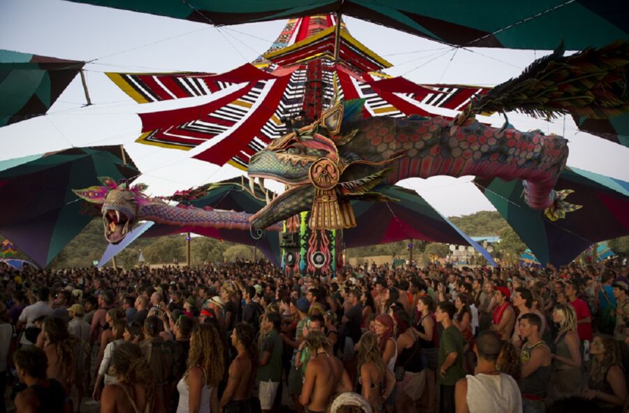 Vibrant festival with dragon sculptures, diverse crowd, colorful canopy, and scenic landscape.
