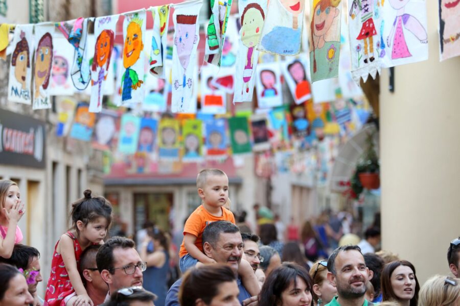 Community event with diverse crowd, childrens artwork, historic town background, and festive atmosphere.