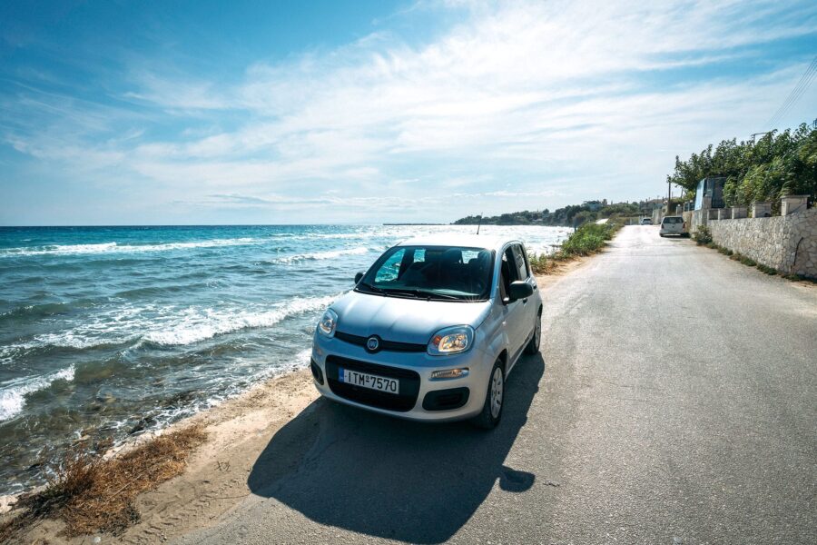 Fiat hatchback by a coastal road, with ocean waves and clear blue sky in Greece.