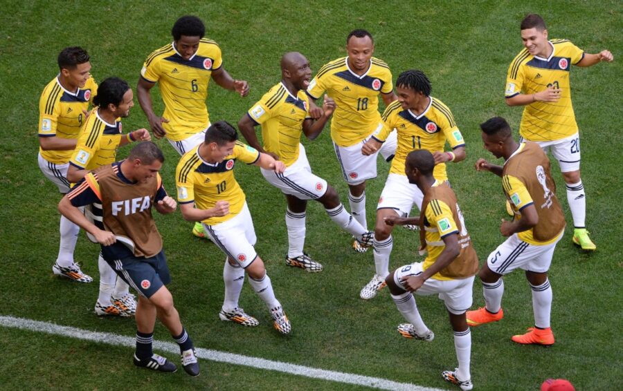 Joyful FIFA Players soccer team in yellow and white uniforms dance in a circle, celebrating on a green field. A coach joins, adding to the festive atmosphere.