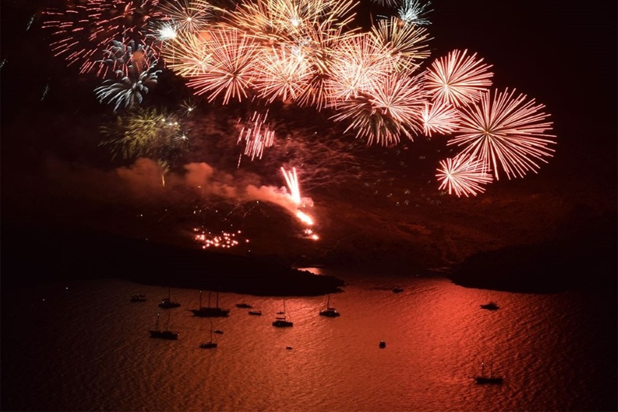 Fireworks display over calm bay, colorful reflections, boats silhouetted, celebrating Ifestia Festival.