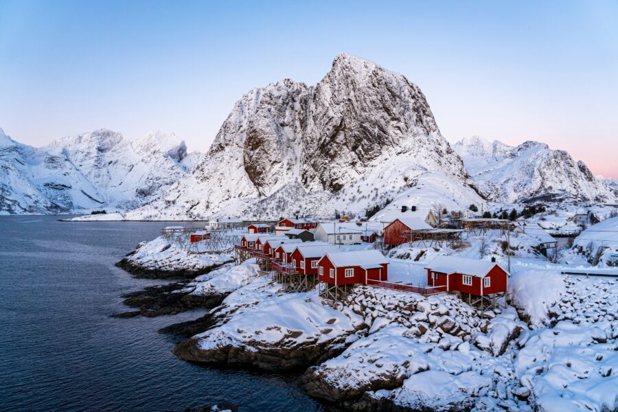 Lofoten, Norway fjord with red cabins on snowy shore beneath towering mountain.