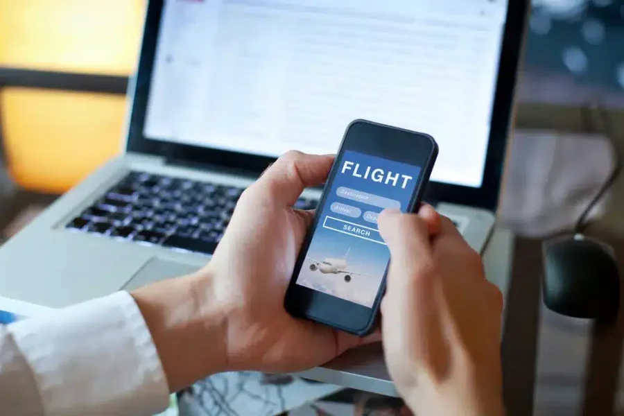 Booking flights on smartphone in professional workspace with laptop in background.