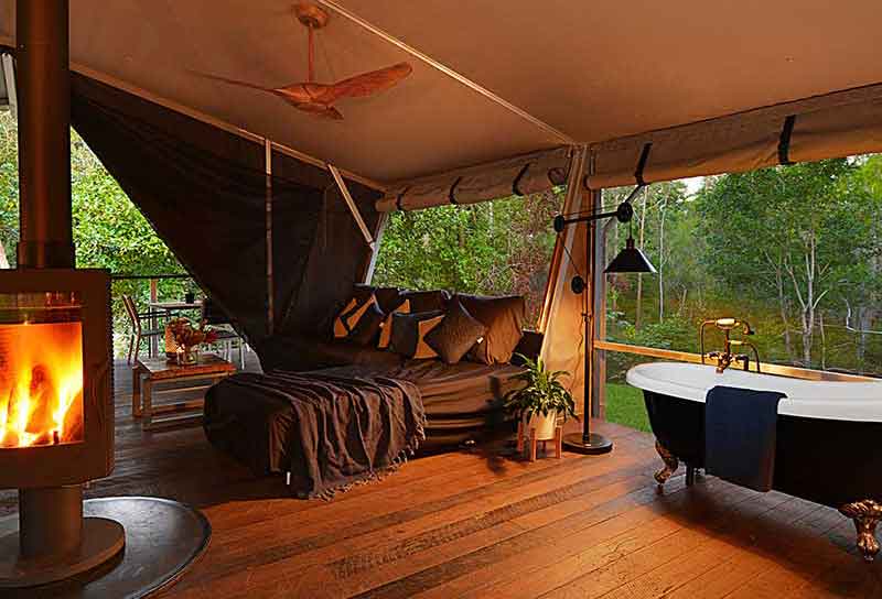 Luxury forest glamping tent interior with wood stove, cozy bed, and freestanding bathtub.