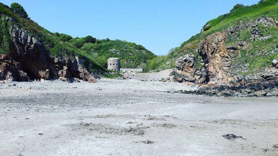 Scenic sandy beach in Forest, Guernsey, with a prominent rock formation rising in the background under sunny weather.

