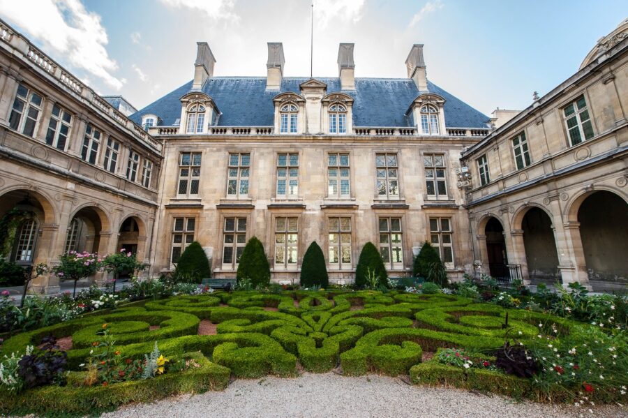 Formal stone courtyard with clipped parterre garden, arcaded wings, and symmetrical Renaissance-style façade.