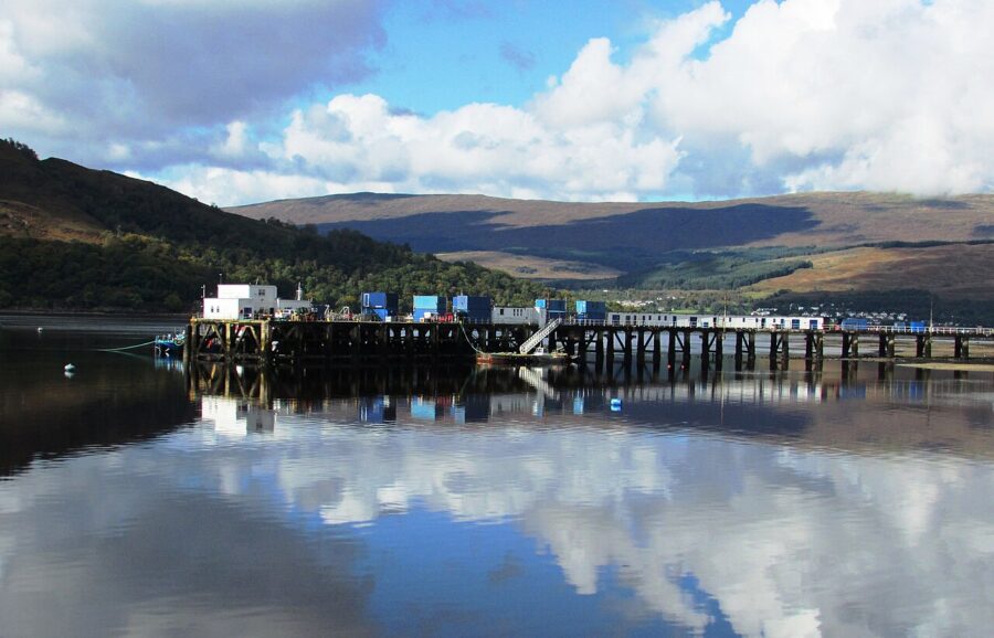 Fort William pier extends into a calm loch, reflecting the sky and clouds. Hills in the background add depth, while the mood is serene and picturesque.