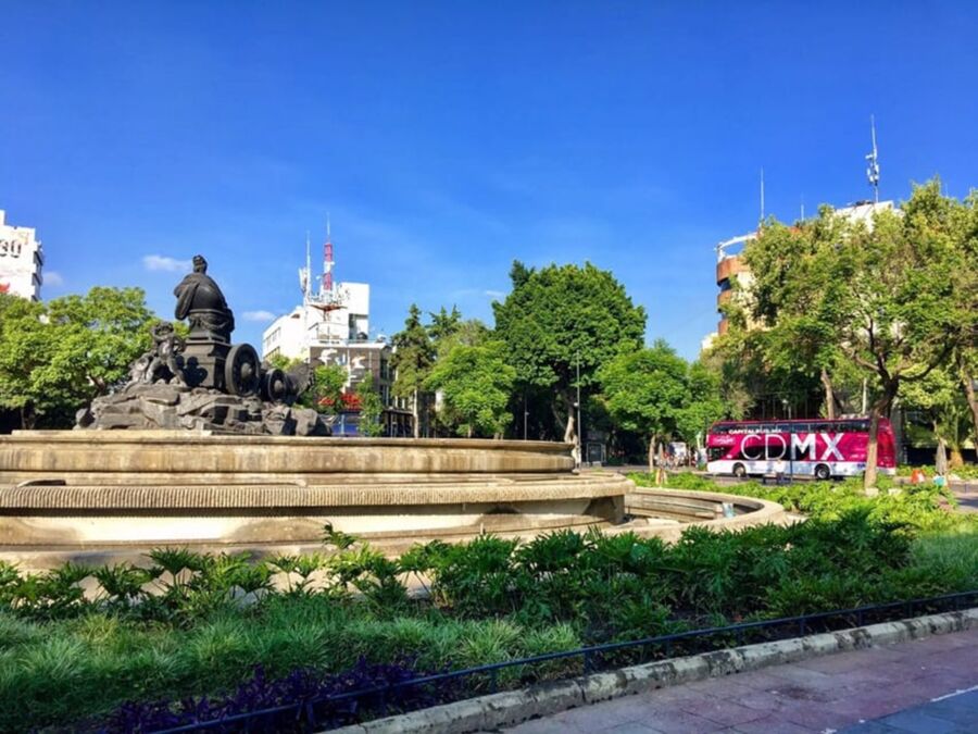 Urban scene in Mexico City featuring a historic fountain, lush greenery, and modern architecture.