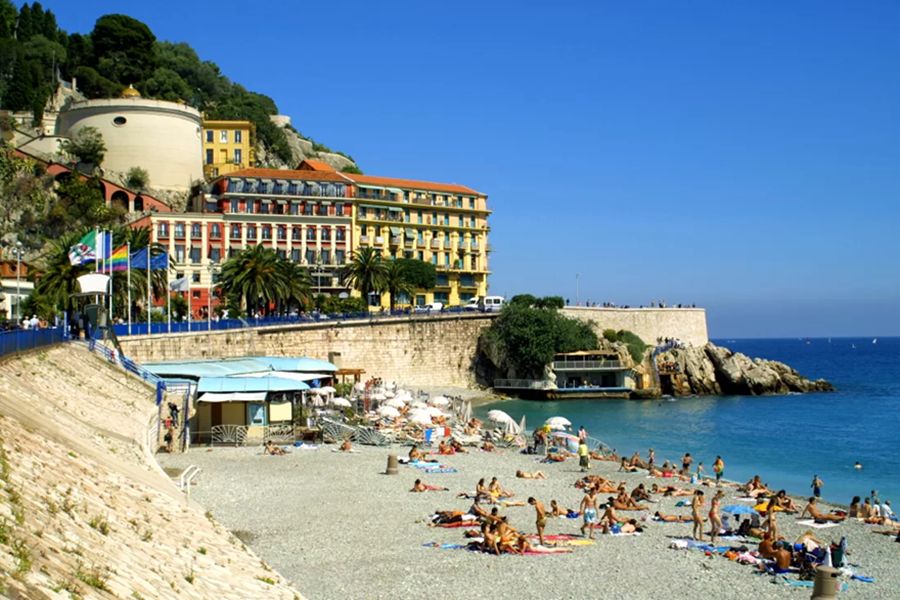 Beautiful coastal beach scene with sunbathers, palm trees, resort, and sailboats on the horizon.