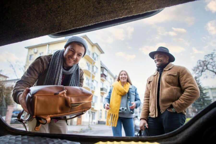 Friends smiling with luggage in an open car trunk on a chilly day.