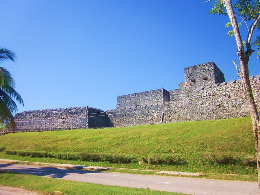 The stone wall of Fuerte de San Felipe Bacalar stands tall, with a tree in front, blending history and natural beauty.