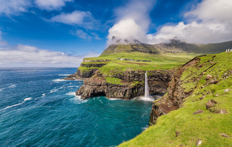 Scenic view of Gasadalur village in the Faroe Islands, featuring dramatic cliffs and a waterfall cascading into the ocean.
