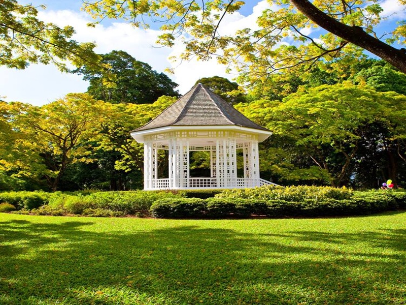 White gazebo in lush park, green lawns, colorful shrubs, trees, sunny sky.