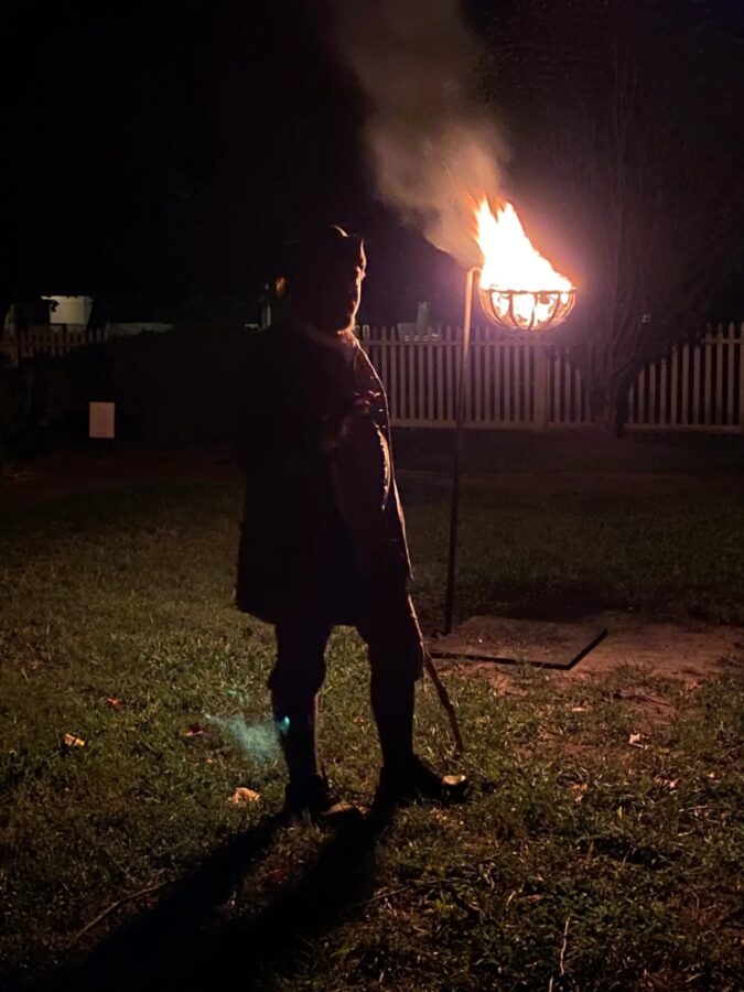 Man with a torch stands in front of a fire, part of the Ghost Stories of Williamsburg guided tour experience.