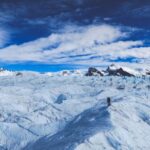 Glacier landscape with undulating ice, mountains, blue sky, and solitary figure walking.