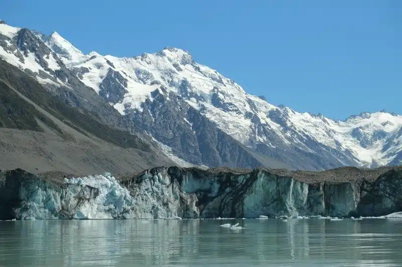 Glacial lake with jagged Tasman Glacier and snow-capped Southern Alps mountains under clear blue sky.