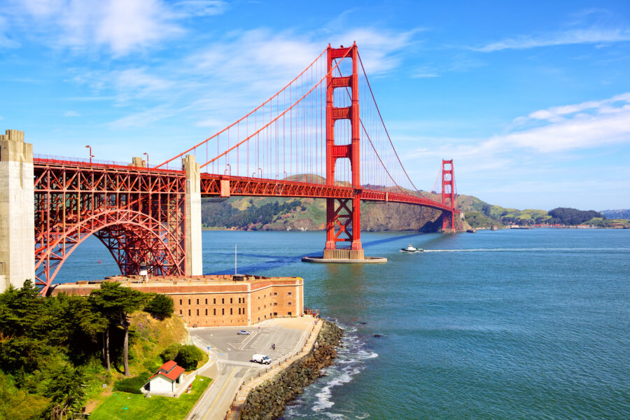 Aerial view of the Golden Gate Bridge under a clear blue sky, spanning over calm waters. Green hills and part of Fort Point are visible below.