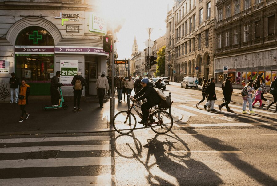 Golden hour city scene with pedestrians, cyclist, scooter, pharmacy, and historic architecture.