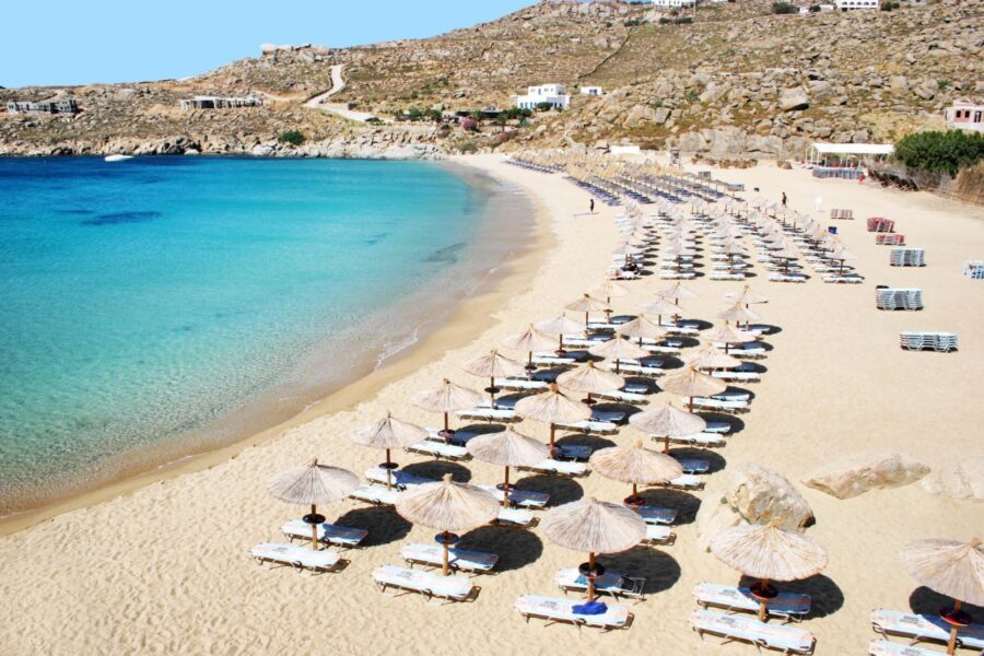 Tranquil beach scene with golden sand, turquoise water, straw umbrellas, and rocky hills.