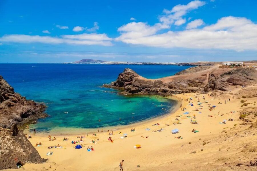 Golden sand beach in Lanzarote with turquoise bay, volcanic cliffs, and relaxing sunbathers.