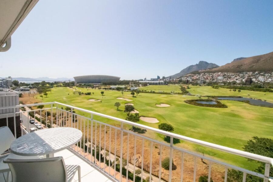 View from balcony: golf course, stadium, city skyline, mountains, and water under a sunny sky.