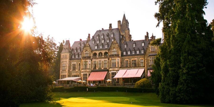 Majestic stone hotel with Gothic architecture, manicured lawn, and golf course amidst lush greenery.