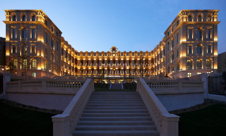Illuminated grand hotel with classic-modern architecture and landscaped entrance under twilight sky.