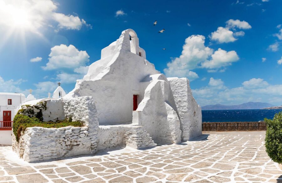 Whitewashed Mediterranean building by tranquil sea with stone path, blue sky, and green vegetation.